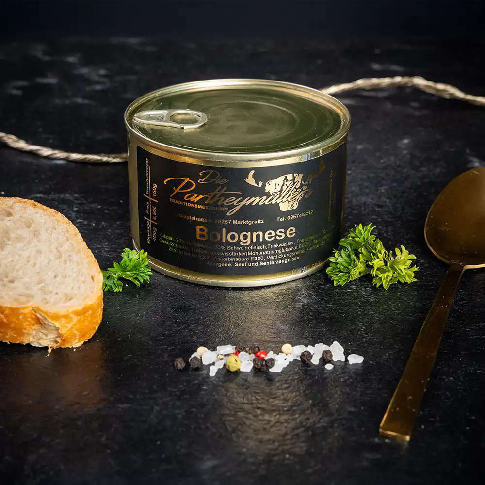 Kleine goldene Konservendose der Metzgerei Partheymüller mit Bolognese-Fleischsauce, edel präsentiert auf dunklem Untergrund mit frischen Kräutern, Pfefferkörnern, Meersalz und einer Baguettescheibe mit Dill.