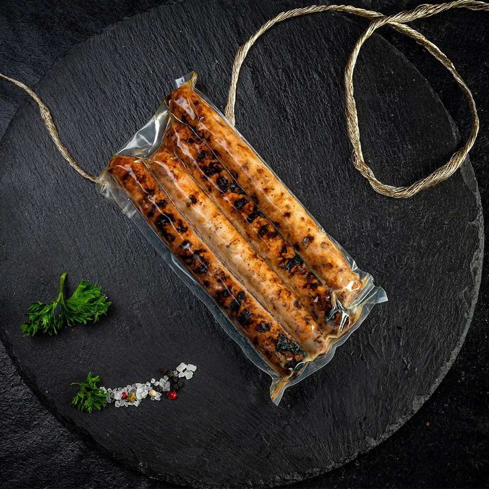 Vier gegrillte Rostbratwürste in Vakuumverpackung, angerichtet auf dunklem Untergrund mit Petersilie, Meersalz und Pfefferkörnern als Dekoration
