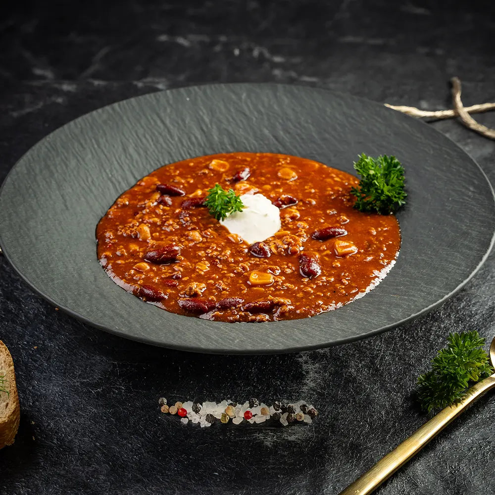 Teller mit würzigem Chili con Carne aus Hackfleisch, Kidneybohnen und Mais, serviert mit einem Klecks Sauerrahm und frischer Petersilie in dunklem tiefen Teller und auf dunklem Untergrund.