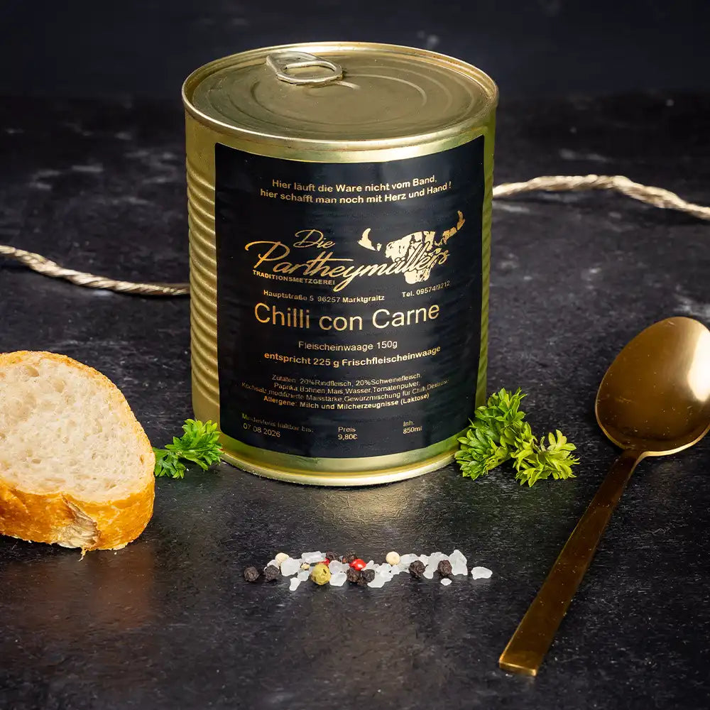 Goldene Konservendose der Metzgerei Partheymüller mit Chili con Carne, edel präsentiert auf dunklem Untergrund mit frischen Kräutern, Pfefferkörnern, Meersalz und einer Baguettescheibe