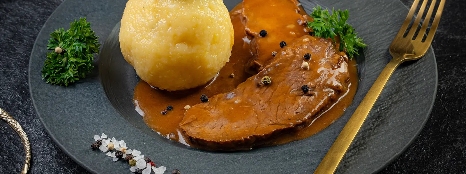 Zwei Scheiben Sauerbraten in Soße neben einem Kloß auf einem dunklen Teller, garniert mit Petersilie. Daneben eine goldene Gabel.