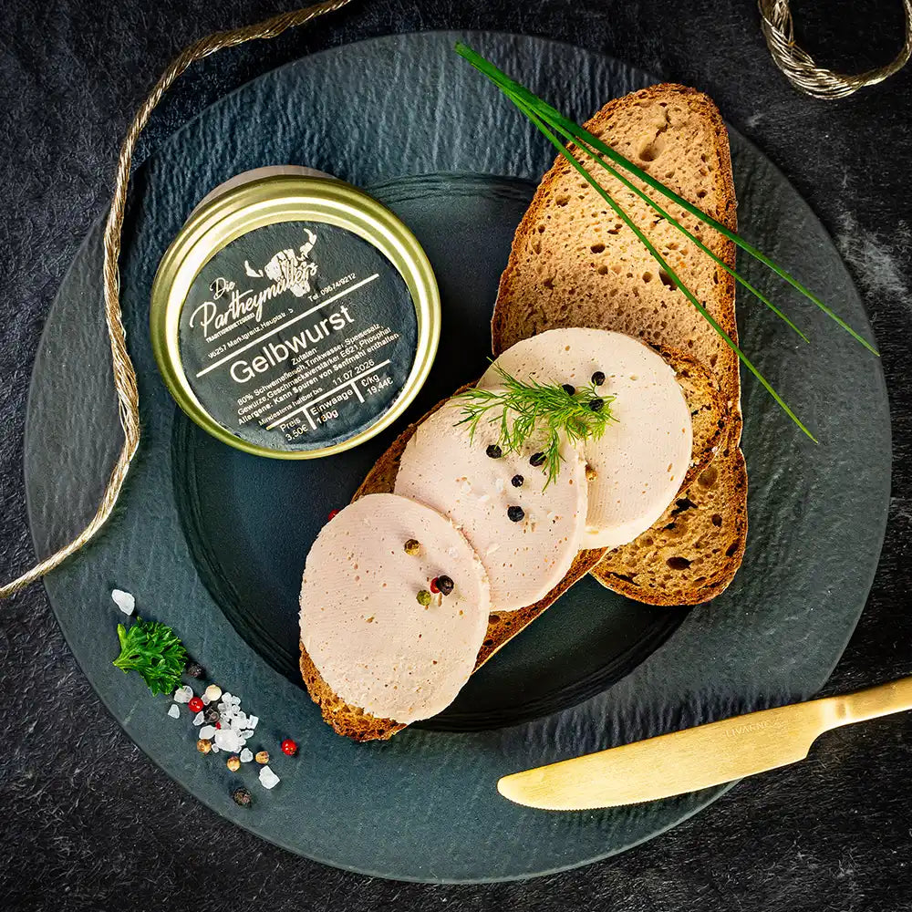 Drei Scheiben Gelbwurst auf Bauernbrot, garniert mit Dill, Schnittlauch und Pfefferkörnern, daneben ein Glas „Gelbwurst“ von Partheymüller auf schwarzem Teller mit goldfarbenem Messer und Gewürzen auf dunklem Untergrund.