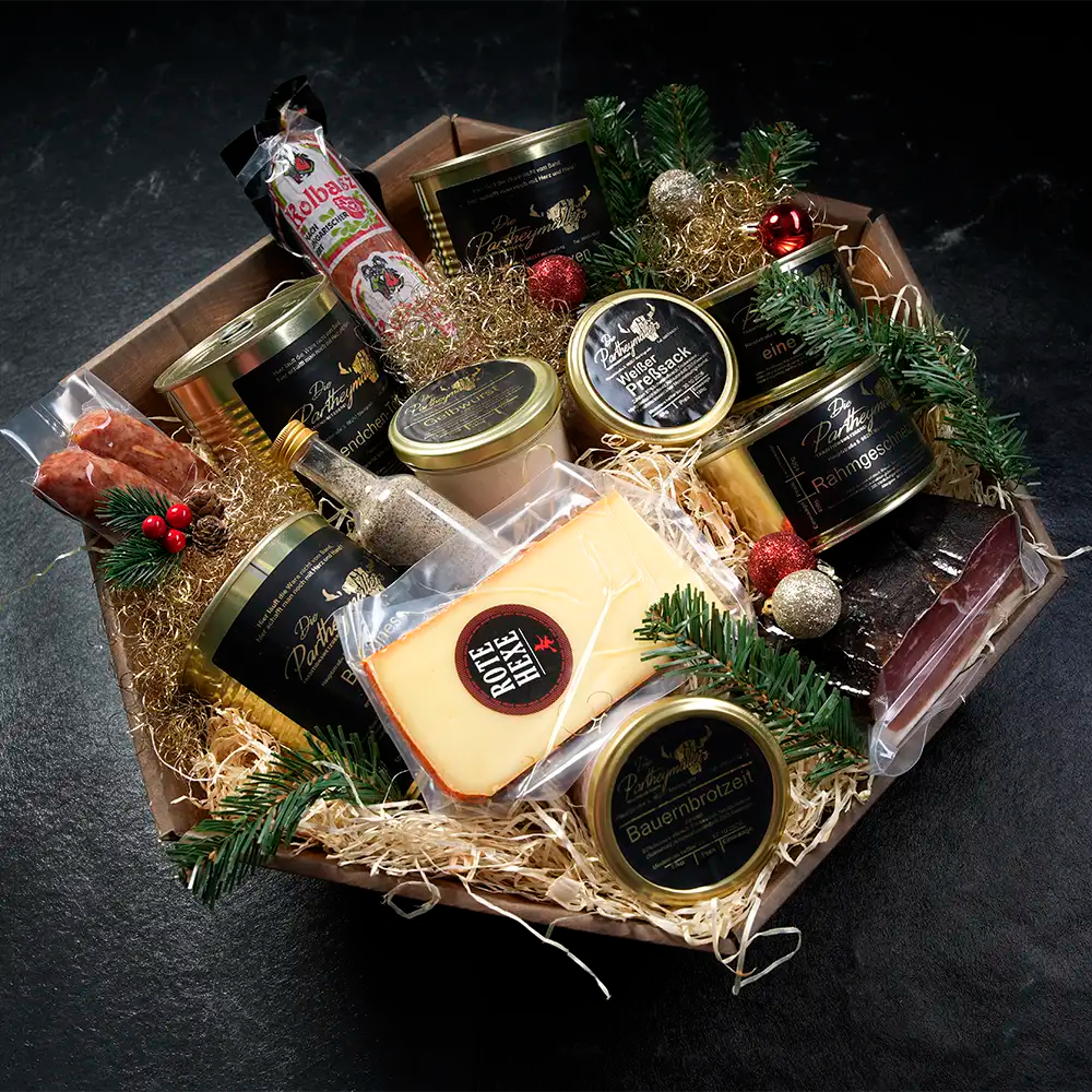 Verschiedene Wurstgläser, Dosen mit schwarzem Etikett, ein Stück Käse, eine Flasche mit Gewürzen und weitere Spezialitäten liegen in einem Holzkorb auf Stroh gebettet vor dunklem Hintergrund.