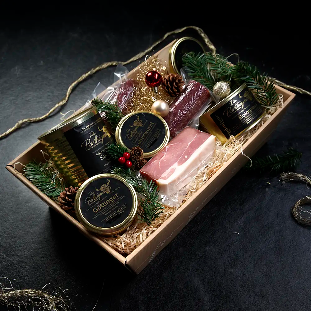 Ein Stück Schinken und verschiedene Dosen mit schwarzem Etikett liegen in einer Holzschachtel mit Stroh ausgebettet vor dunklem Hintergrund.