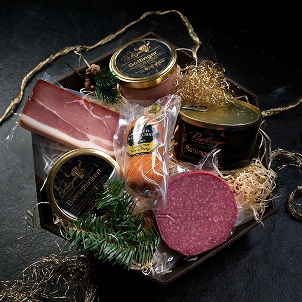 Schachtel mit Wurstdosen, Schinken und Salami auf Stroh gebettet vor dunklem Hintegrund.