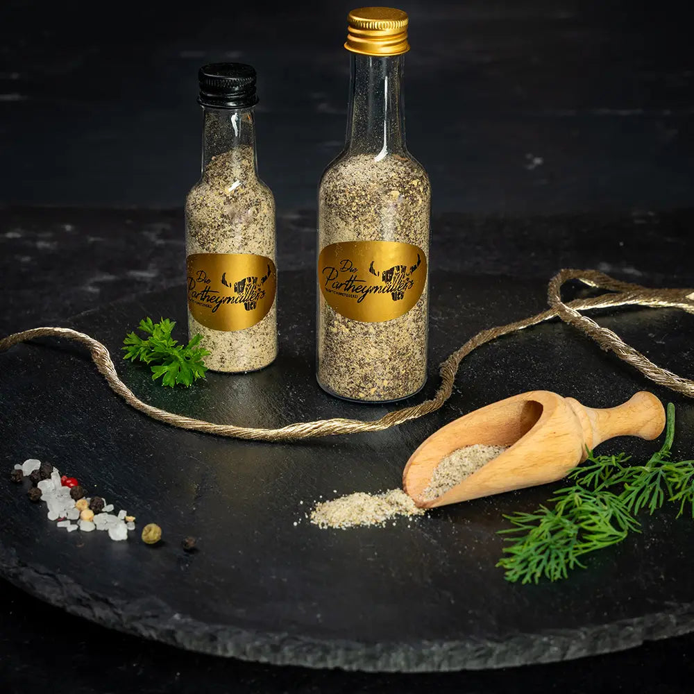 Zwei Gewürzflaschen mit goldfarbenem Etikett der Metzgerei Partheymüller auf schwarzer Steinplatte, daneben Holzschaufel mit ausgeschüttetem Gewürz, Pfefferkörner, grobes Salz, Petersilie und Dillzweig auf dunklem Untergrund.