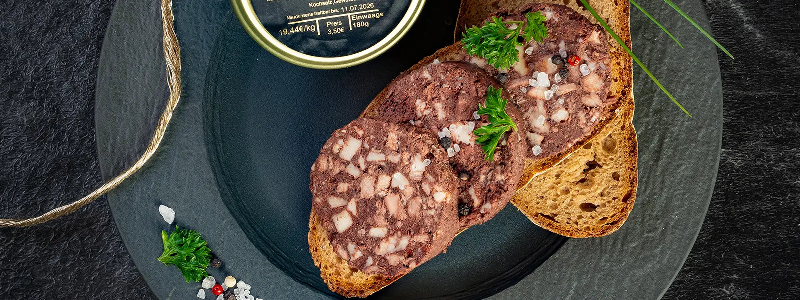 Blutwurst auf einer Scheibe Brot angerichtet mit Petersilie neben einem Wurstglas auf einem dunklen Teller.