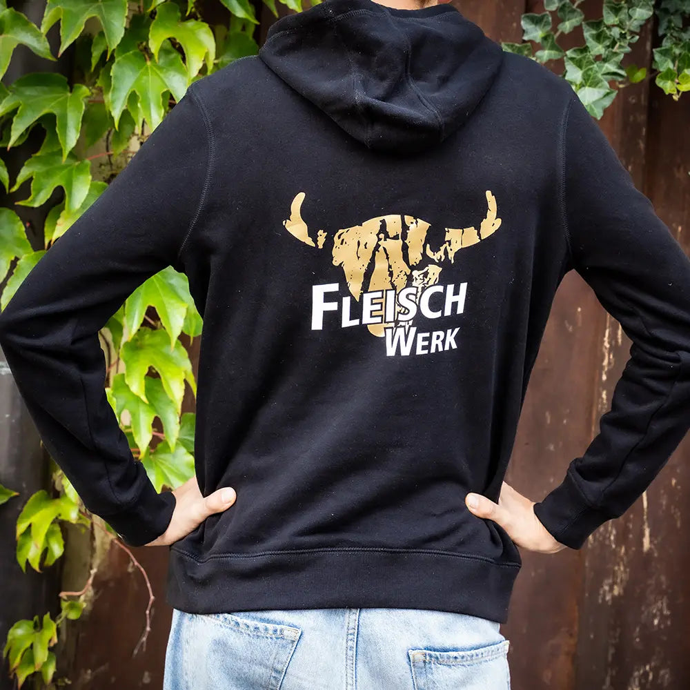 Schwarzer Hoodie mit gold-weißem „FleischWerk“-Logo und stilisiertem Rinderkopf auf dem Rücken, getragen von einer Person vor einer Holzwand mit Efeu und grünen Blättern.