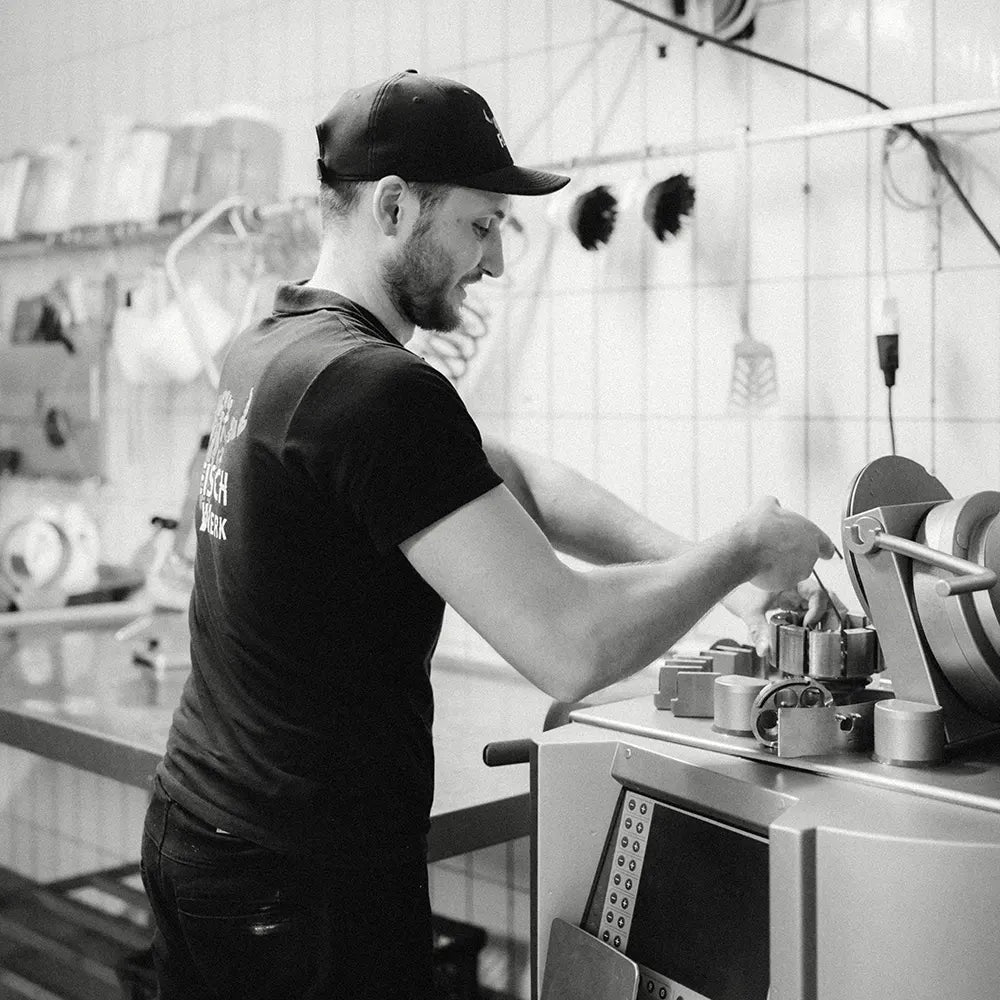 Mann mit in schwarzem Shirt und schwarzer Cap arbeitet an einer Fleischverarbeitungsmaschine in einem gekachelten Produktionsraum