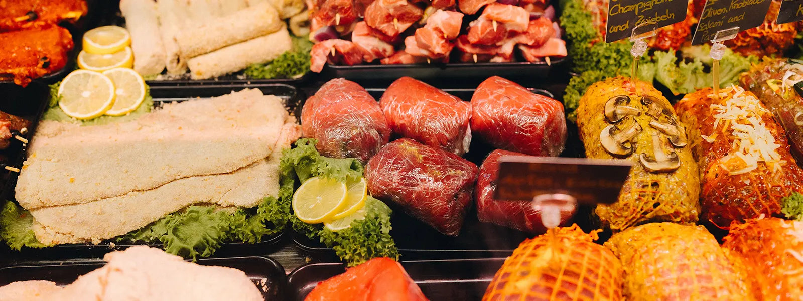 Vielfältige Fleischtheke mit panierten Schnitzeln, gefüllten Rouladen in Frischhaltefolie, Fleischspießen und dekorierten Braten mit Pilzen und Käse, angerichtet auf Salatblättern und garniert mit Zitronenscheiben