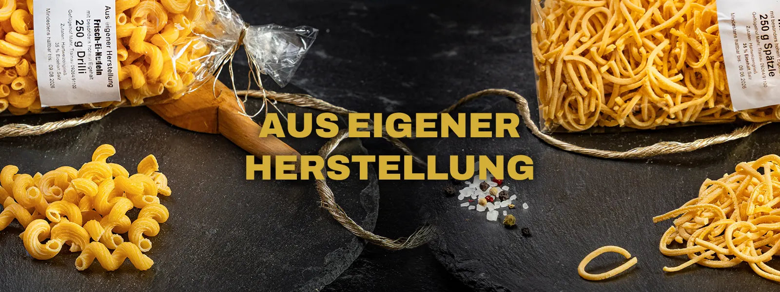 Zwei Sorten Nudeln in Klarsichtverpackung mit Etikett „Aus eigener Herstellung“ – links Drilli-Nudeln, rechts Spätzle – angerichtet auf schwarzem Schieferuntergrund mit Gewürzen und goldgelber Aufschrift in der Mitte: „Aus eigener Herstellung"