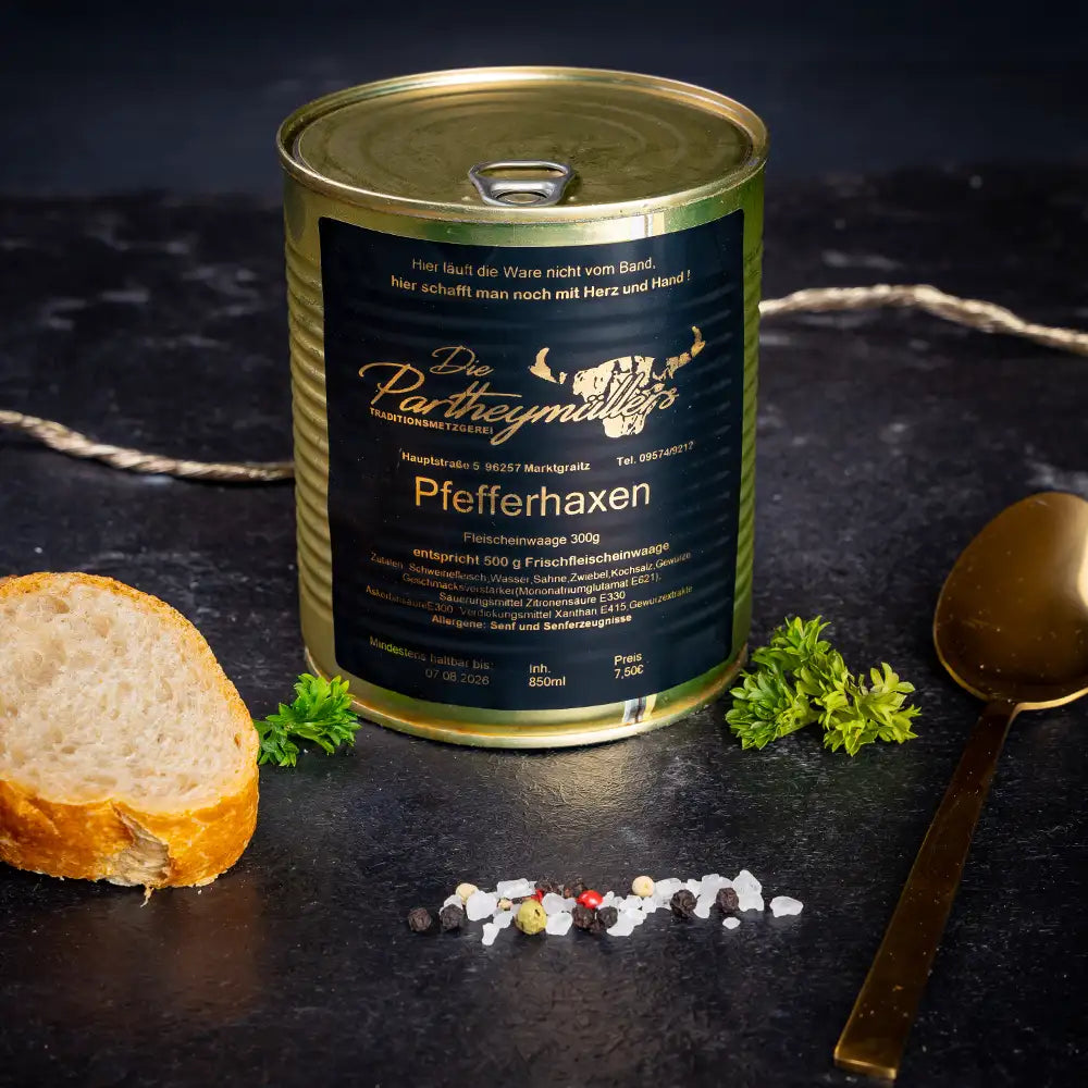 Goldene Konservendose der Metzgerei Partheymüller mit Pfefferhaxe edel präsentiert auf dunklem Untergrund mit goldenem Löffel, frischen Kräutern, Pfefferkörnern, Meersalz und einer Baguettescheibe