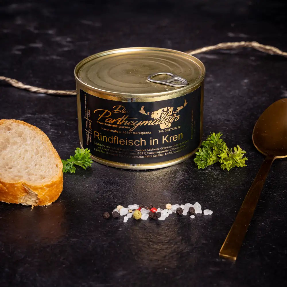 Kleine goldene Konservendose der Metzgerei Partheymüller mit Rindfleisch in Kren edel präsentiert auf dunklem Untergrund mit goldenem Löffel, frischen Kräutern, Pfefferkörnern, Meersalz und einer Baguettescheibe.