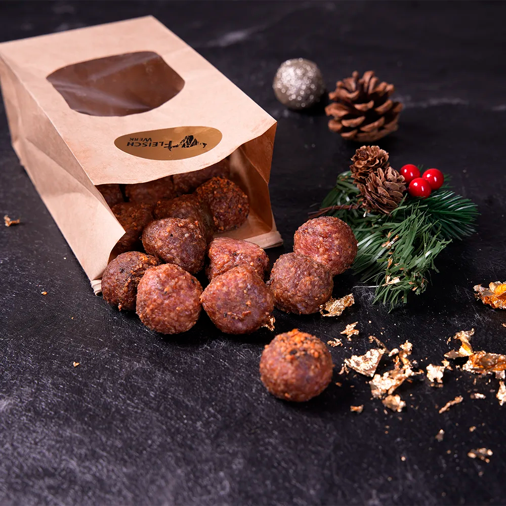 Mehrere Salamikugeln rollen aus kartonfarbenem umgefallenen Tütchen vor dunklem Hintergrund.