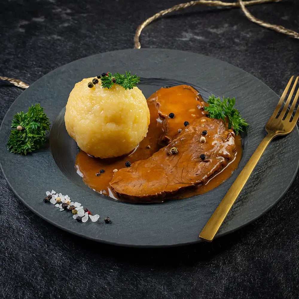 Sauerbraten mit zwei Scheiben Fleisch in dunkler Sauce und einem Kartoffelknödel auf schwarzem Teller, garniert mit Petersilie, Pfefferkörnern und grobem Salz, daneben eine goldene Gabel auf dunklem Untergrund