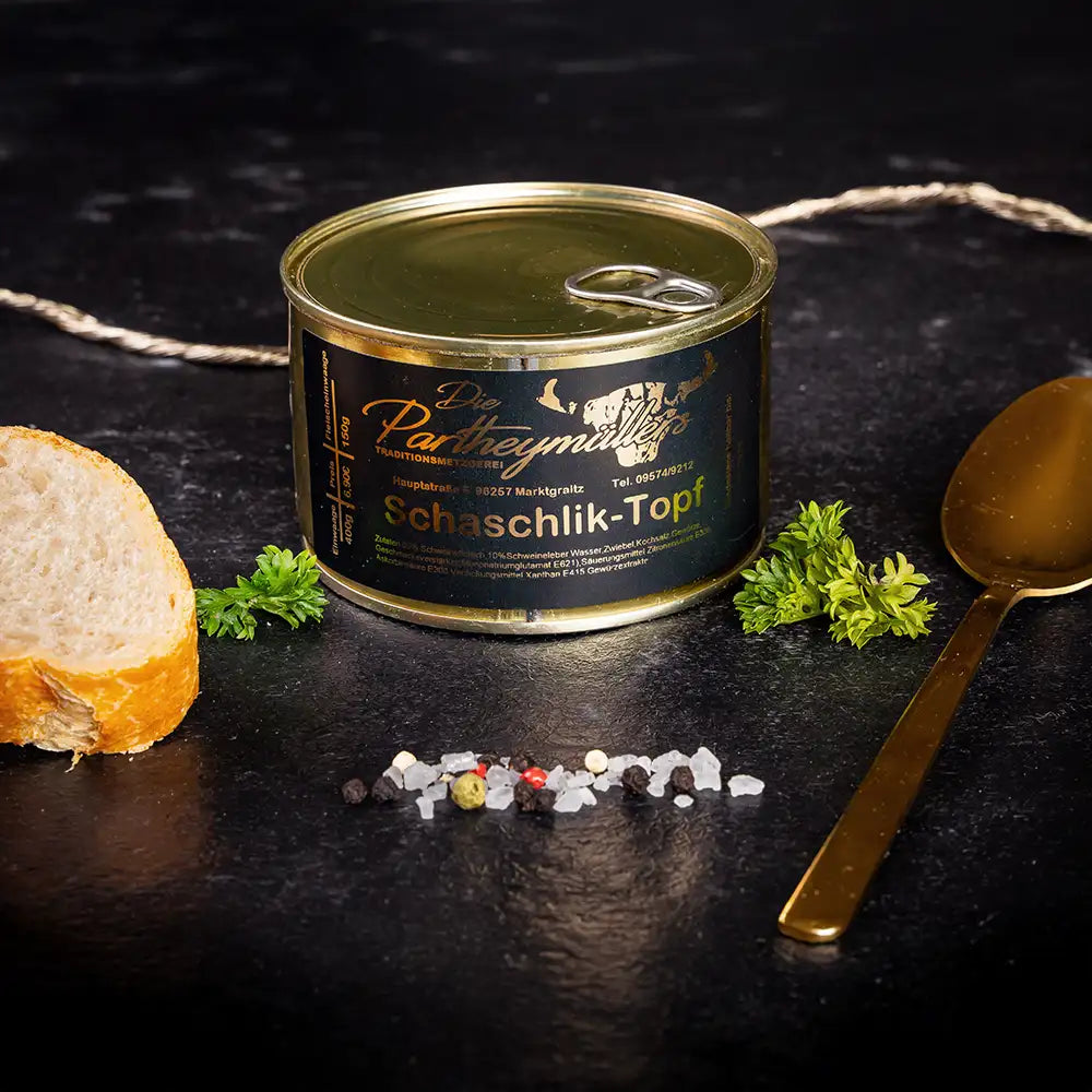 Kleine goldene Konservendose der Metzgerei Partheymüller mit Schaschlik-Topf edel präsentiert auf dunklem Untergrund mit goldenem Löffel, frischen Kräutern, Pfefferkörnern, Meersalz und einer Baguettescheibe