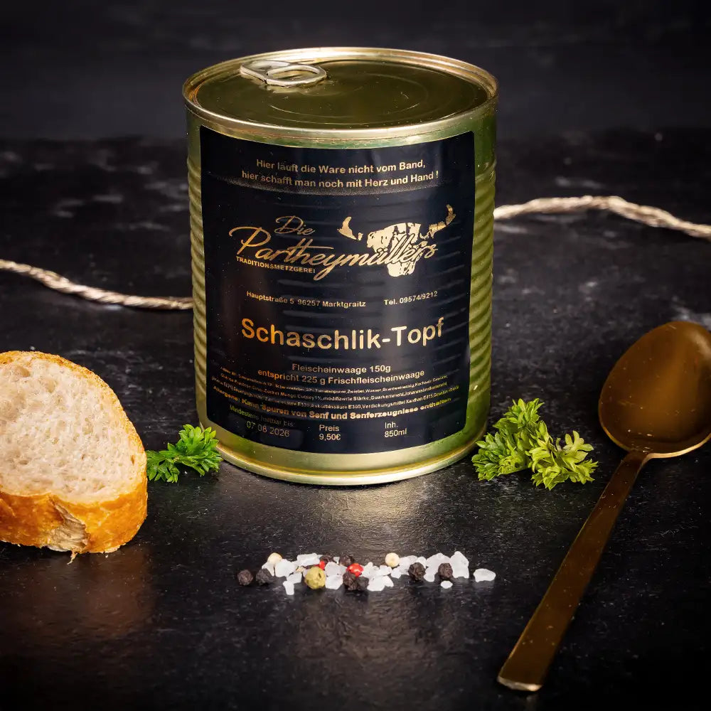 Goldene Konservendose der Metzgerei Partheymüller mit Schaschlik-Topf edel präsentiert auf dunklem Untergrund mit goldenem Löffel, frischen Kräutern, Pfefferkörnern, Meersalz und einer Baguettescheibe