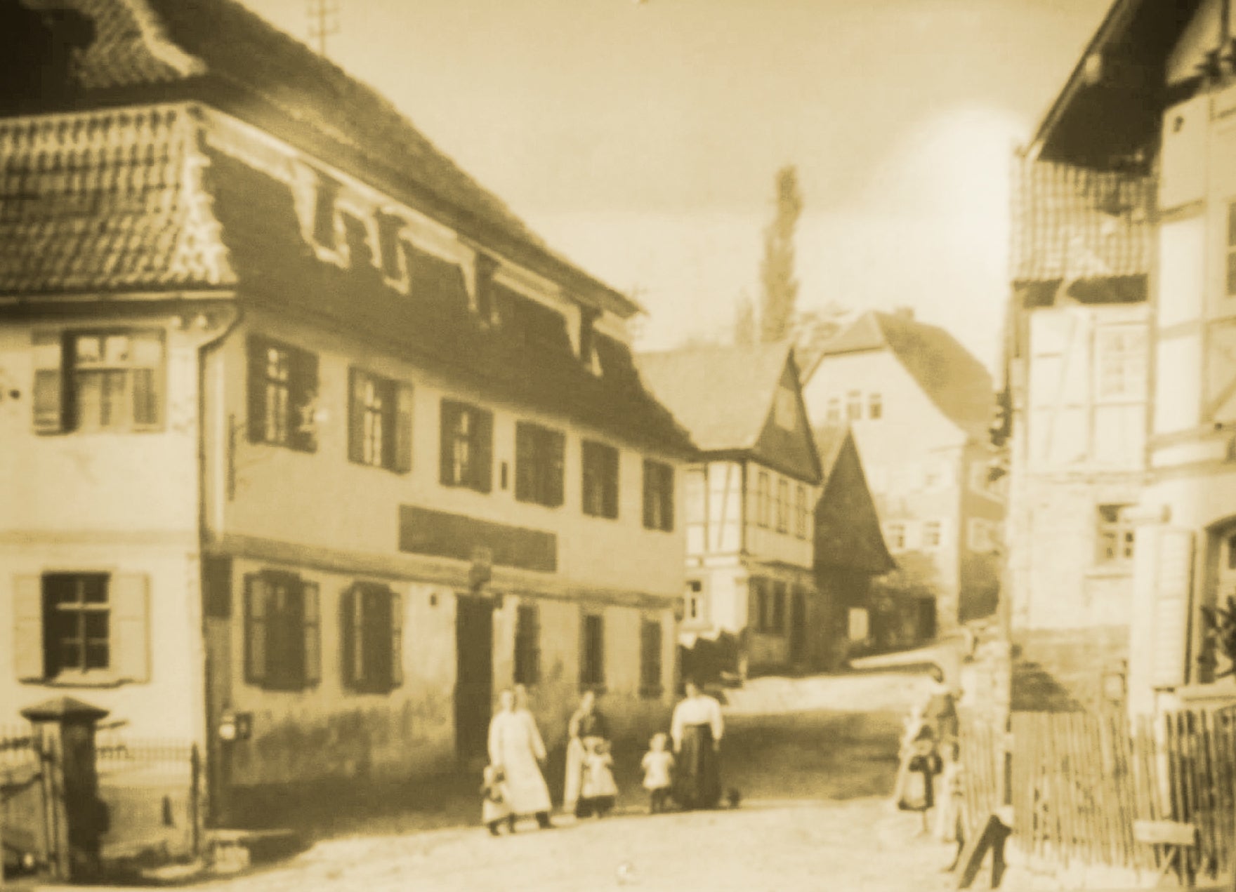 Historische Fotografie einer alten Dorfstraße mit Fachwerkhäusern und mehreren Personen in traditioneller Kleidung, darunter Frauen und Kinder; das Bild ist in Sepia gehalten und vermittelt eine Szenerie aus dem frühen 20. Jahrhundert