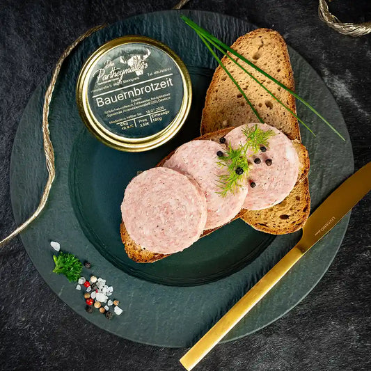 Drei Scheiben Bauernbrotzeit auf einer Scheibe Bauernbrot, daneben eine weitere Scheibe Brot, angerichtet mit Pfefferkörnern und frischem Dill, daneben ein Glas „Bauernbrotzeit“ von der Marke Partheymüller auf einem dunklen Teller mit einem goldenem Messer.