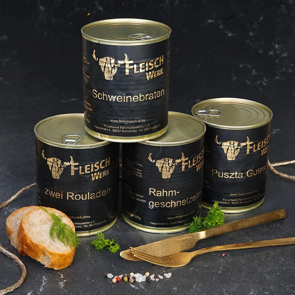 Vier Dosen mit dunklem Etikett stehen aufeinander gestapelt auf dunkler Schieferplatte. Daneben liegt goldenes Besteck und Baguettescheiben.