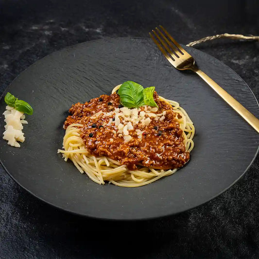 Spaghetti mit herzhafter Bolognese-Sauce, garniert mit geriebenem Parmesan und frischem Basilikum, serviert auf einem schwarzen Teller mit goldener Gabel