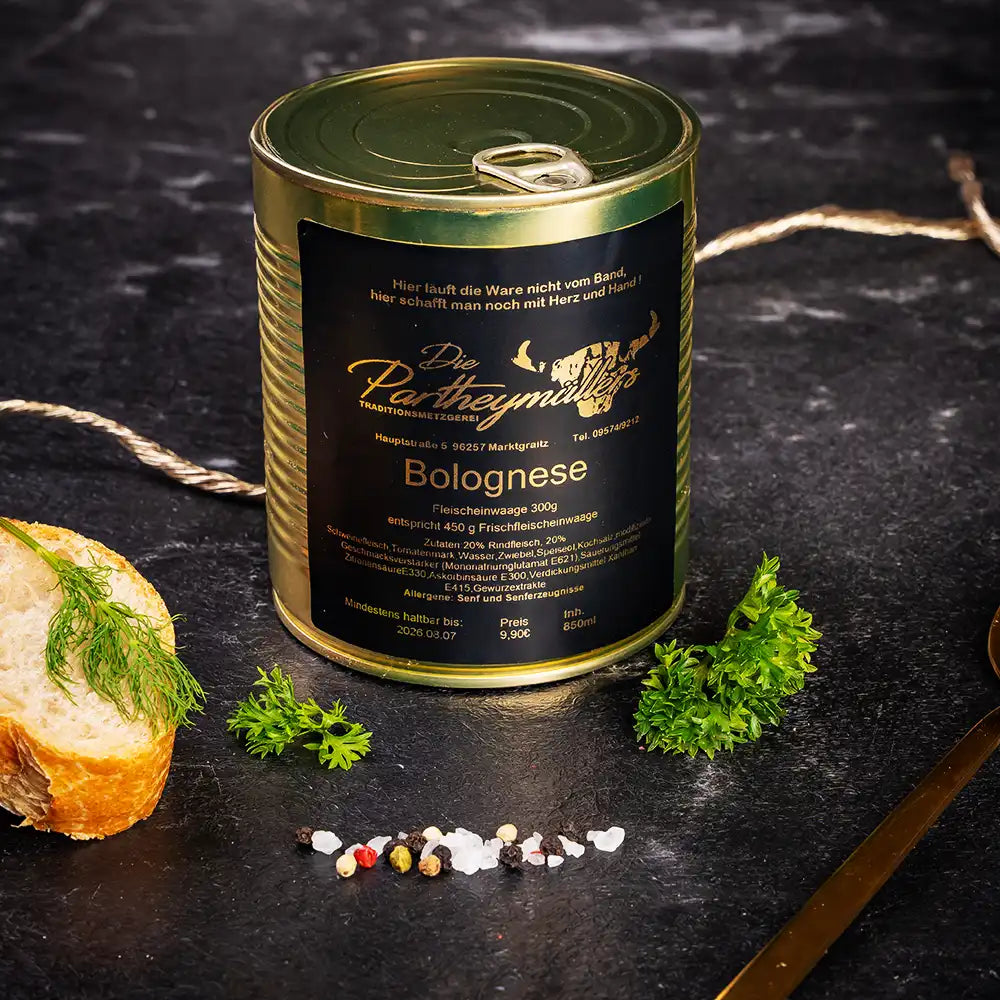 Goldene Konservendose der Metzgerei Partheymüller mit Bolognese-Fleischsauce, edel präsentiert auf dunklem Untergrund mit frischen Kräutern, Pfefferkörnern, Meersalz und einer Baguettescheibe mit Dill.