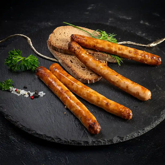 Vier gebratene Rostbratwürste auf dunkler Platte, serviert mit zwei Scheiben Bauernbrot, frischen Kräutern sowie Meersalz und Pfefferkörnern als Garnitur