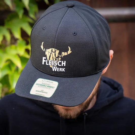Schwarze Cap mit gold-weißem „FleischWerk“-Logo und stilisiertem Rinderkopf, getragen von einer Person in schwarzem Hoodie, Sticker mit Hinweis auf recyceltes Material sichtbar auf dem Schirm