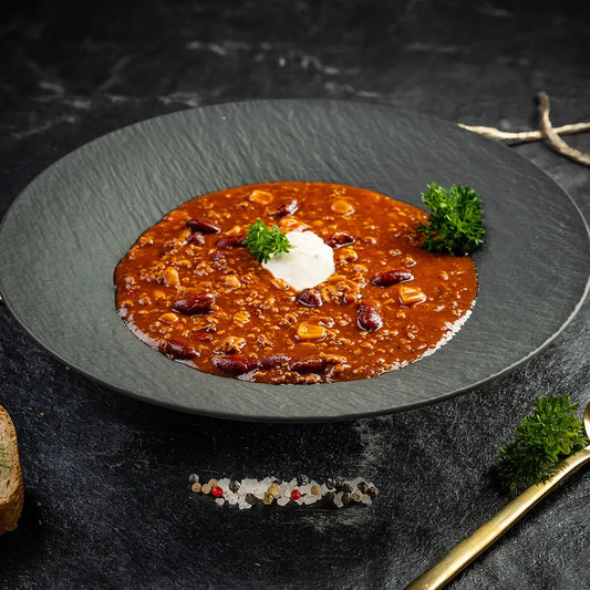 Teller mit würzigem Chili con Carne aus Hackfleisch, Kidneybohnen und Mais, serviert mit einem Klecks Sauerrahm und frischer Petersilie in dunklem tiefen Teller und auf dunklem Untergrund.