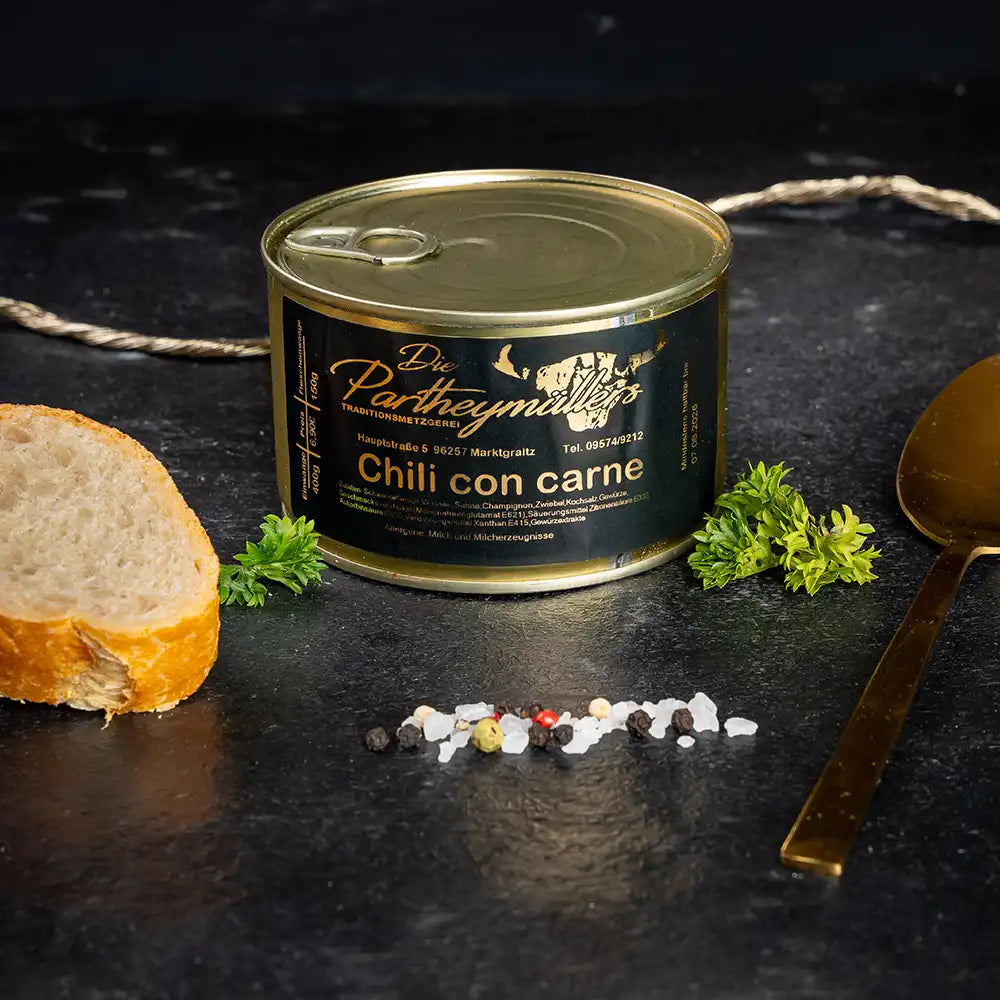 Kleine goldene Konservendose der Metzgerei Partheymüller mit Chili con Carne, edel präsentiert auf dunklem Untergrund mit frischen Kräutern, Pfefferkörnern, Meersalz und einer Baguettescheibe