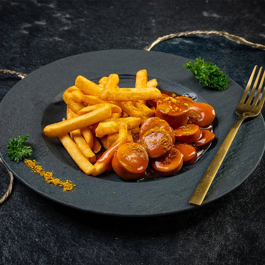 Portion Currywurst mit würziger Currysauce und goldbraunen Pommes frites, angerichtet auf einem schwarzen Teller mit goldener Gabel und frischer Petersilie auf dunklem Untergrund