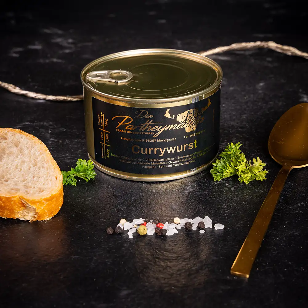 Kleine goldene Konservendose der Metzgerei Partheymüller mit Currywurst, edel präsentiert auf dunklem Untergrund mit frischen Kräutern, Pfefferkörnern, Meersalz und einer Baguettescheibe