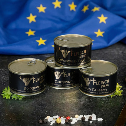 Vier Dosen mit dunklem Etikett stehen aufeinander gestapelt auf dunkler Schieferplatte vor einer Europa Flagge.
