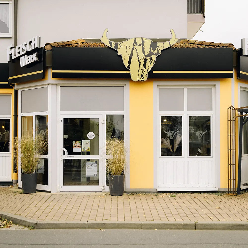 Außenansicht eines Ladengeschäfts mit gelber Fassade, schwarzem Vordach und großem Rinderkopf-Logo über dem Eingang. Links und rechts vom Eingang stehen hohe, schwarze Pflanzkübel mit Ziergräsern. Aufschrift über dem Laden: „Fleisch Werk“.