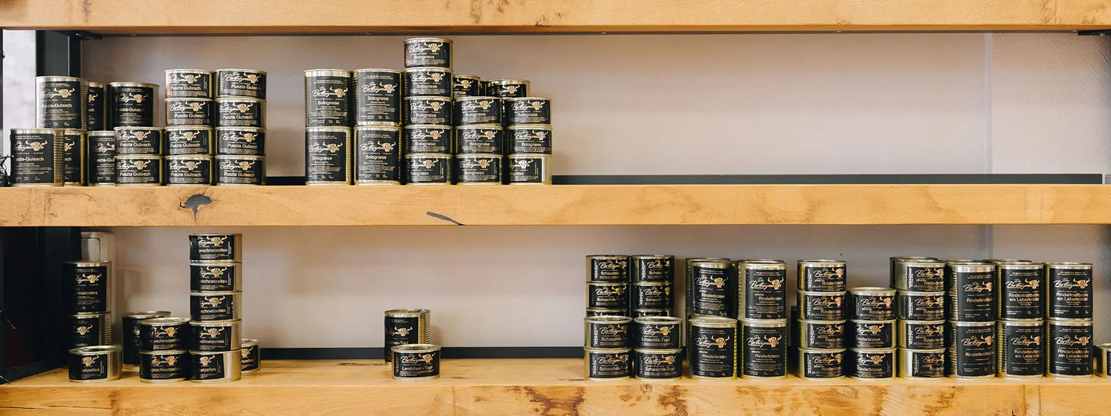 Regal mit gestapelten schwarz-goldenen Dosen verschiedener Wurst- und Fleischspezialitäten der Marke Partheymüller, sortiert nach Sorten und Größen, präsentiert auf zwei Holzablagen vor hellem Hintergrund.