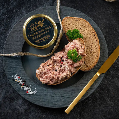 Dunkler, runder Teller mit zwei Scheiben Bauernbrot, eine davon dick mit Fränkischem Aufstrich „Frankenschmaus“ bestrichen und mit Petersilie sowie Pfefferkörnern garniert; daneben eine Glas„Frankenschmaus“ von Partheymüller, ein goldenes Messer und etwas grobes Salz mit Pfefferkörnern und einer goldenen Kordel.