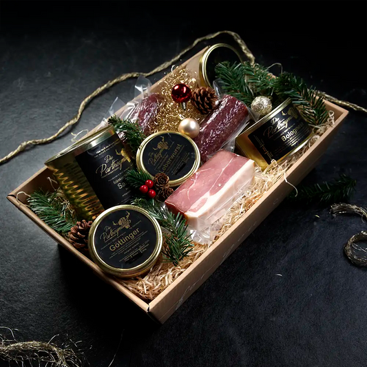 Ein Stück Schinken und verschiedene Dosen mit schwarzem Etikett liegen in einer Holzschachtel mit Stroh ausgebettet vor dunklem Hintergrund.
