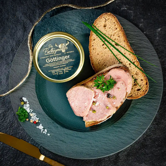 Aufschnittscheiben der Goettinger Wurst auf Bauernbrot, garniert mit Petersilie, Pfefferkörnern und Schnittlauch, daneben ein Glas Goettinger von der Metzgerei Partheymüller auf einem dunklen Teller mit Gewürzen daneben ein goldenes Messer.