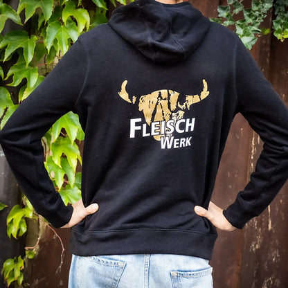 Schwarzer Hoodie mit gold-weißem „FleischWerk“-Logo und stilisiertem Rinderkopf auf dem Rücken, getragen von einer Person vor einer Holzwand mit Efeu und grünen Blättern.