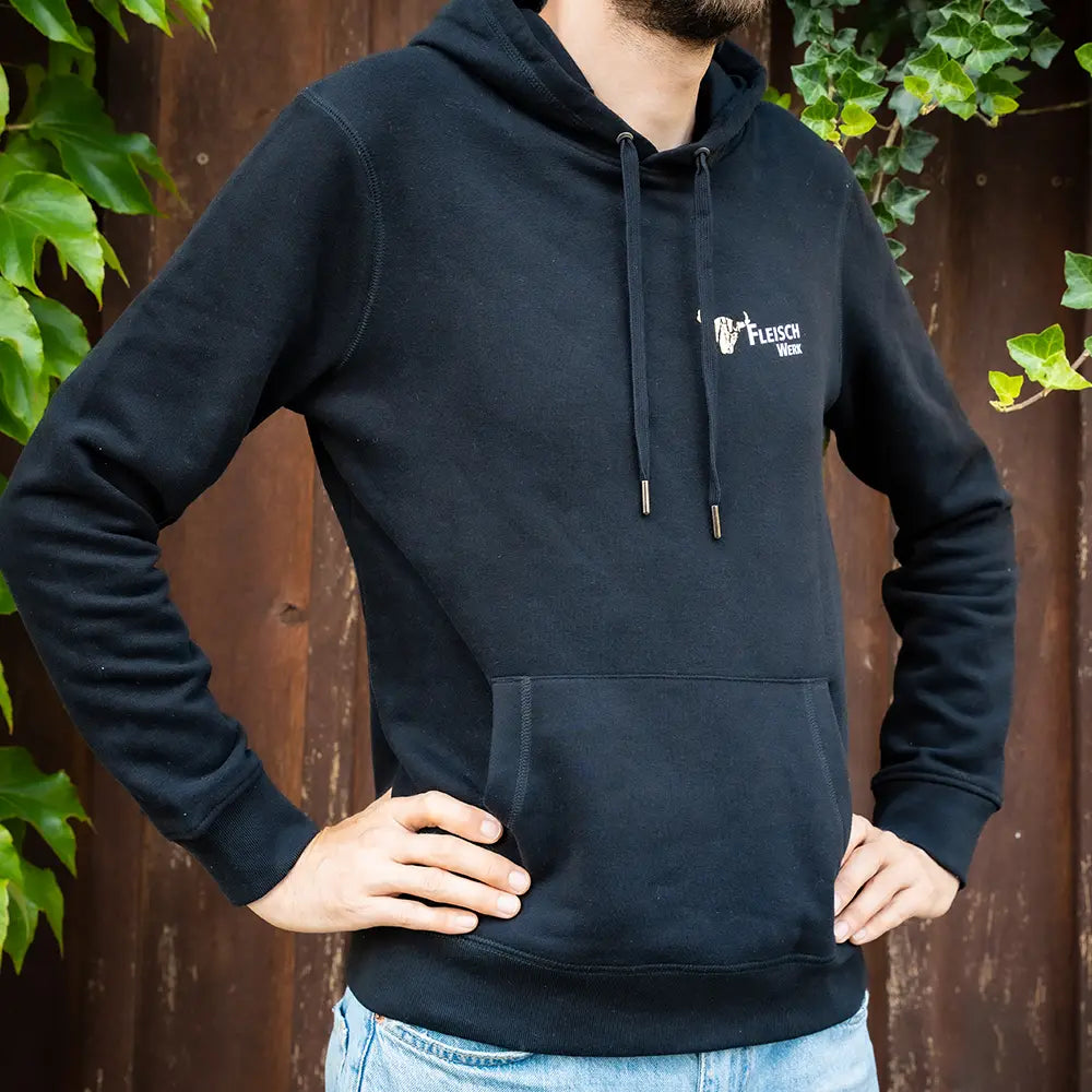 Schwarzer Hoodie mit „FleischWerk“-Logo auf der Brust, getragen von einer Person mit Jeans vor einer Holzwand mit grünem Efeu
