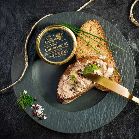 Hausgemachte Leberwurst der Metzgerei Partheymüller auf einer Scheibe Bauernbrot, darunter eine zweite Scheibe Brot, verziert mit Pfefferkörnern, Dill und Schnittlauch, daneben das Glas Leberwurst auf einem dunklen Teller mit goldfarbenem Messer und Gewürzen