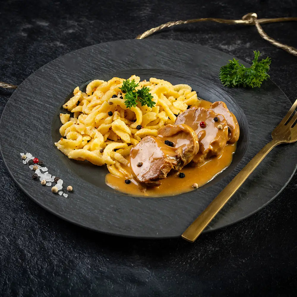 Spätzle mit zarten Lendchen in Rahmsauce, serviert auf einem dunklen Teller mit goldener Gabel, garniert mit frischer Petersilie und Gewürzen.