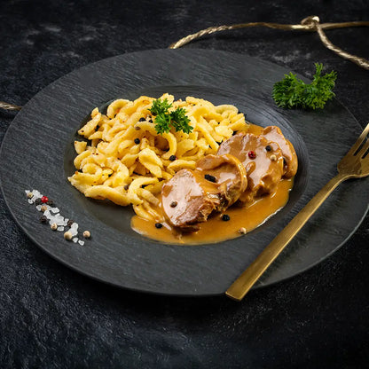 Spätzle mit zarten Lendchen in Rahmsauce, serviert auf einem dunklen Teller mit goldener Gabel, garniert mit frischer Petersilie und Gewürzen.