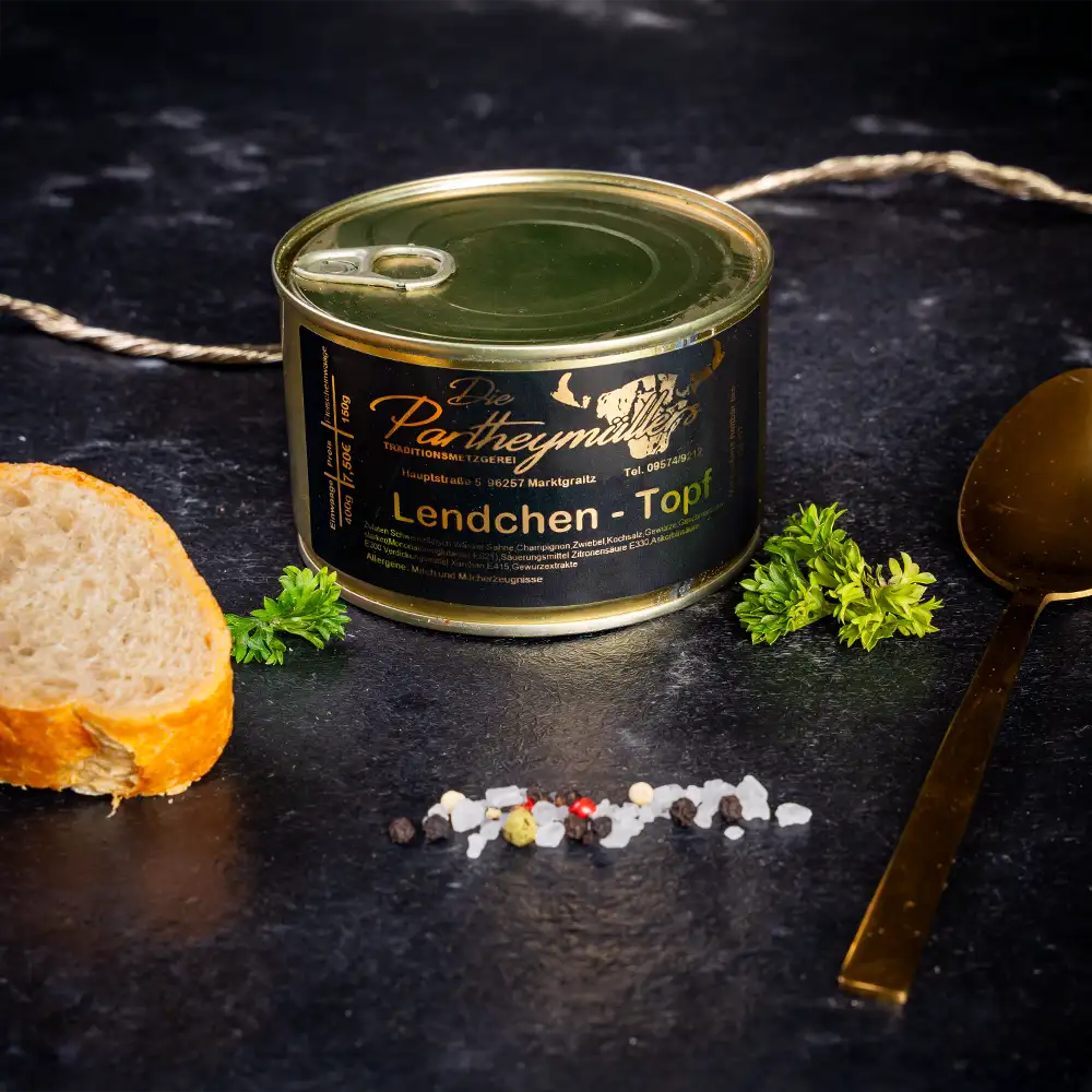 Kleine goldene Konservendose der Metzgerei Partheymüller mit Lendchentopf, edel präsentiert auf dunklem Untergrund mit frischen Kräutern, Pfefferkörnern, Meersalz und einer Baguettescheibe.