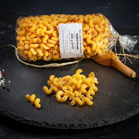 Eine durchsichtige Verpackung mit Drilli-Nudeln aus Hartweizengrieß mit weißem Etikett liegend auf dunkler Steinplatte, daneben lose Nudeln, Holzlöffel, Gewürze und eine goldfarbene Kordel auf dunklem Untergrund.