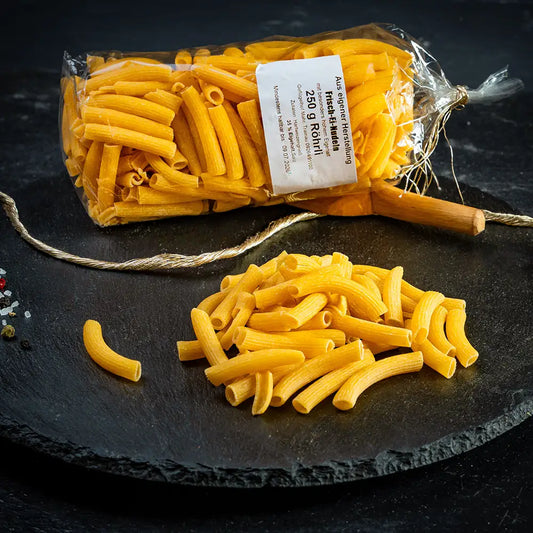Eine durchsichtige Verpackung mit Roehrli-Nudeln aus Hartweizengrieß mit weißem Etikett liegend auf dunkler Steinplatte, daneben lose Nudeln, Holzlöffel, Gewürze und eine goldfarbene Kordel auf dunklem Untergrund.