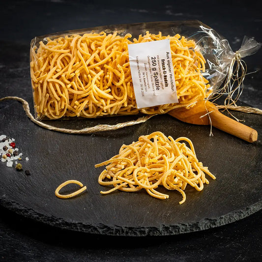 Eine durchsichtige Verpackung mit Spätzle-Nudeln aus Hartweizengrieß mit weißem Etikett liegend auf dunkler Steinplatte, daneben lose Nudeln, Holzlöffel, Gewürze und eine goldfarbene Kordel auf dunklem Untergrund.