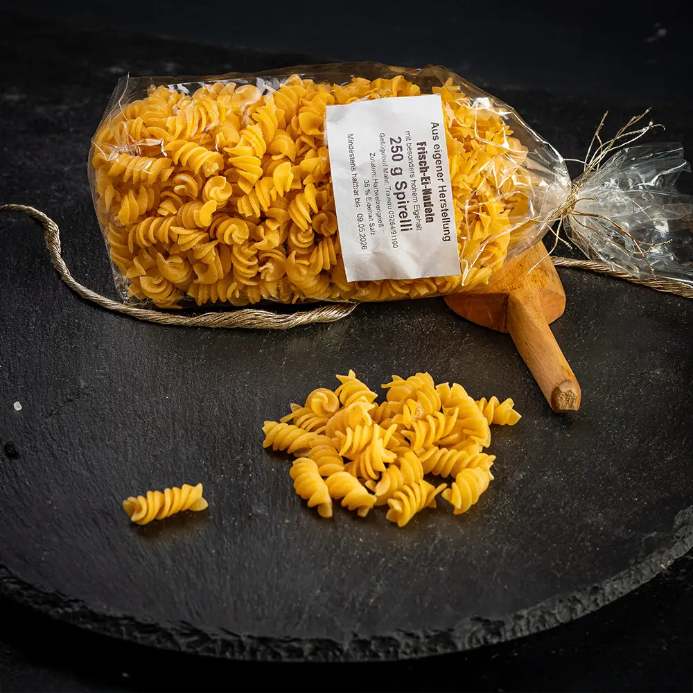Eine durchsichtige Verpackung mit Spirelli-Nudeln aus Hartweizengrieß mit weißem Etikett liegend auf dunkler Steinplatte, daneben lose Nudeln, Holzlöffel, Gewürze und eine goldfarbene Kordel auf dunklem Untergrund.
