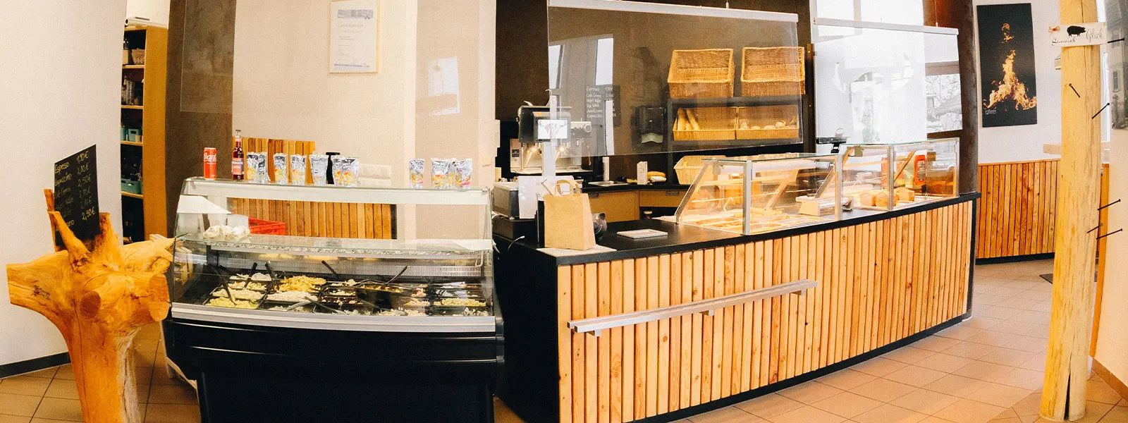Innenansicht einer Metzgerei mit moderner Verkaufstheke aus Holzlamellen, links eine Salattheke mit Getränken und Verpackungen, rechts die Haupttheke mit Wurst- und Fleischwaren, im Hintergrund Brotkörbe und Kaffeemaschine, helle Wände und Fliesenboden