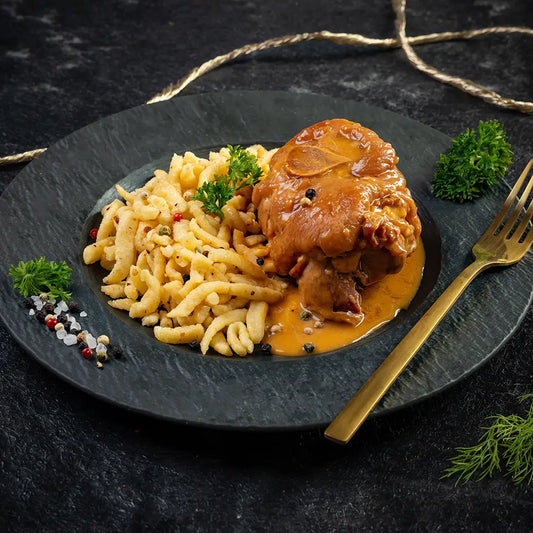 Pfefferhaxe in heller Sauce mit Spätzle auf schwarzem Teller, garniert mit Petersilie, Pfefferkörnern und grobem Salz, daneben eine goldene Gabel und dekorative Kordel auf dunklem Untergrund
